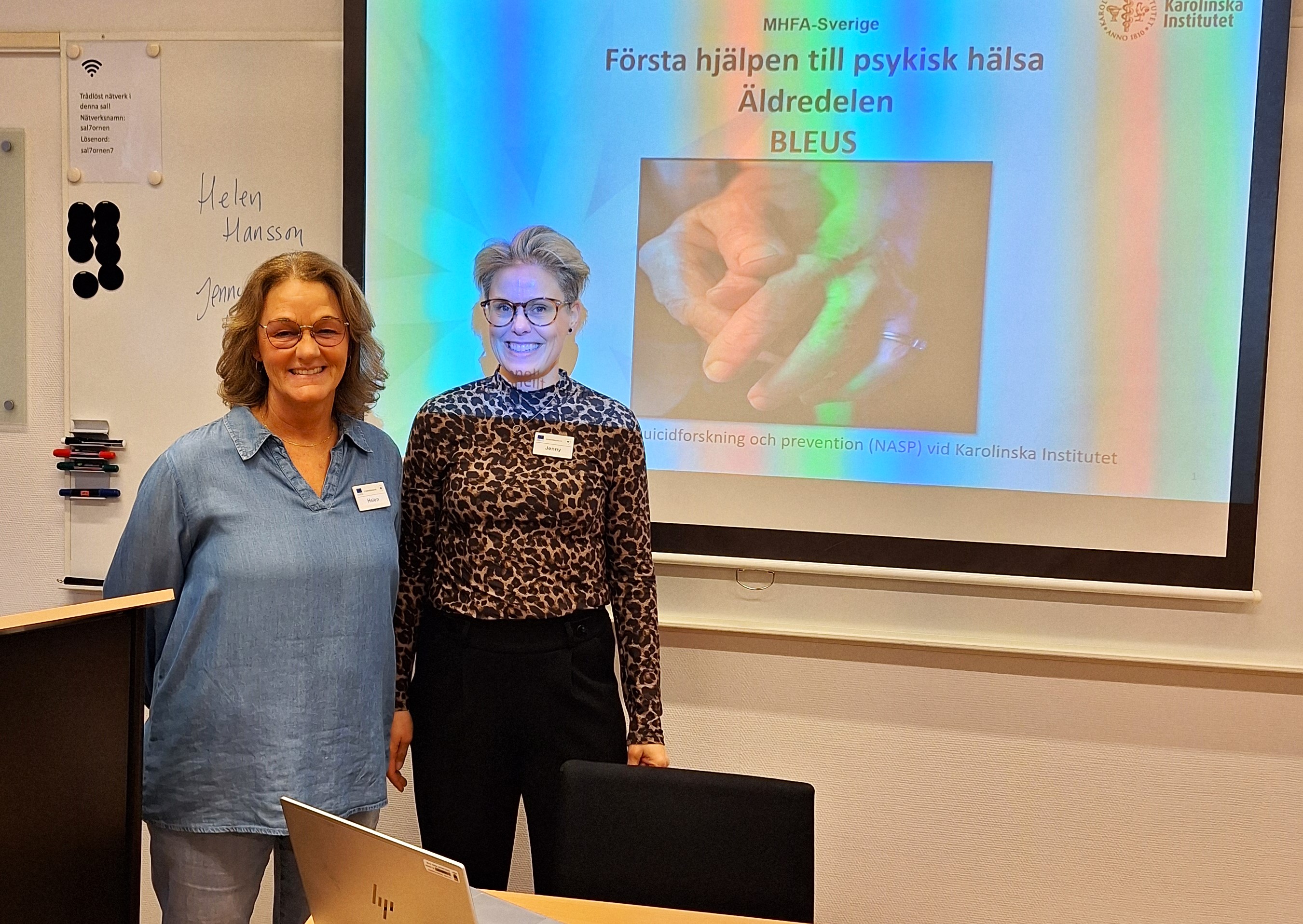 Två leende kvinnor står i en föreläsningssal. Text på skärm MHFA Äldre