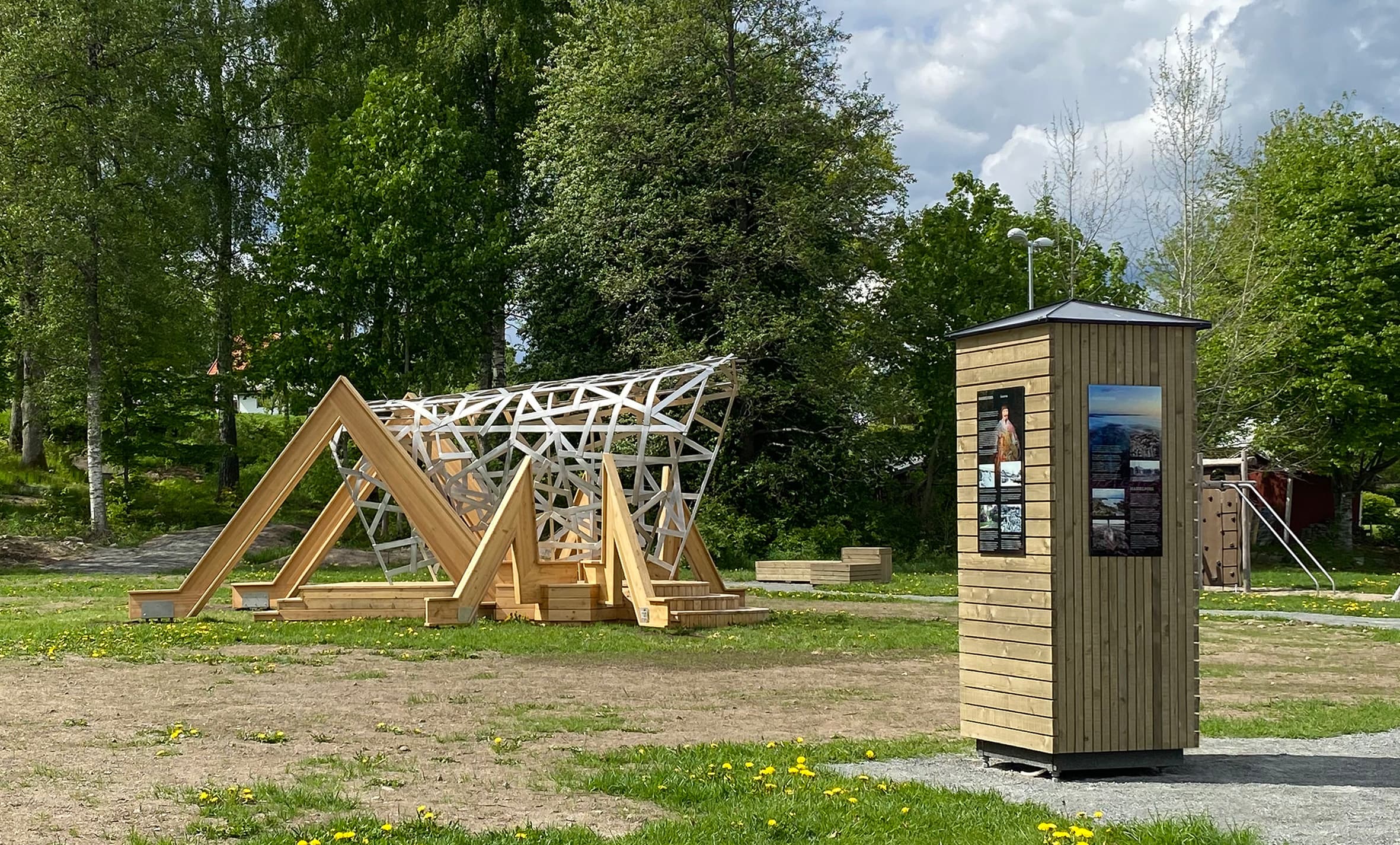 Fotografi av parkmiljö med stor semiabstrakt skulptur av gräshoppa med ben av trä och kropp av ett nät av metall.