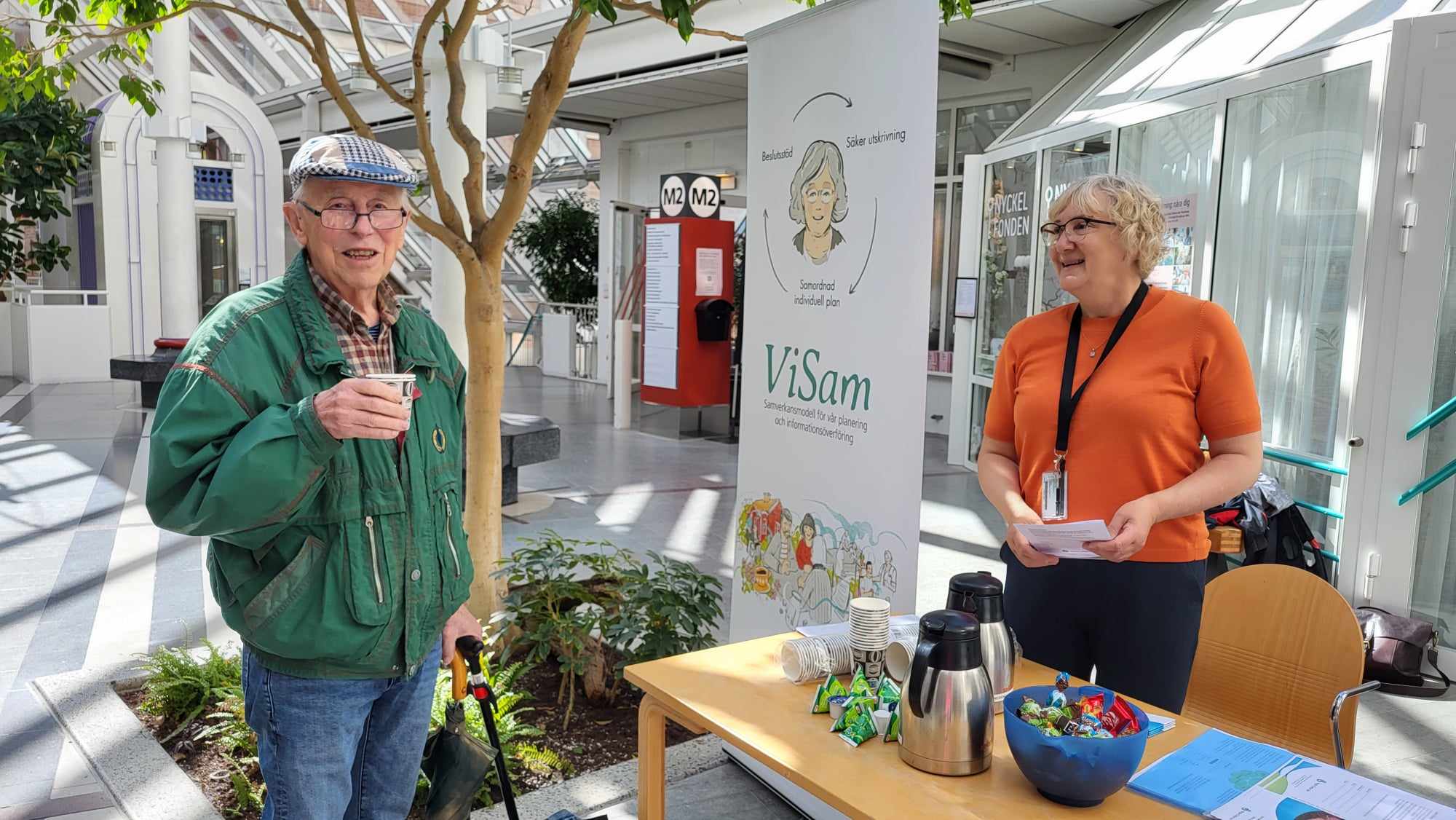 En äldre man i keps dricker kaffe och pratar med en kvinna i orange top.