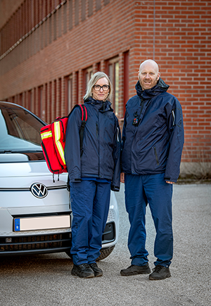 Två personer i vårdkläder står framför en vit bil.