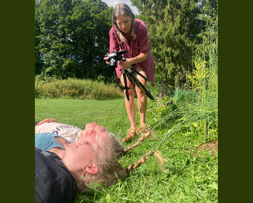 Kvinna fotograferar tre kvinnor som ligger ner med varsin fläta inflätad i högt gräs